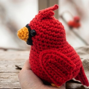 May include: A red crocheted cardinal bird plush toy featuring a bright orange beak, a black mask around the eyes, and a small crest on its head. The bird is crafted with visible yarn stitches and has a rounded body shape with a small tail.