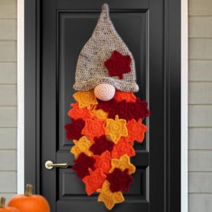 May include: A handmade crochet gnome door decoration with a gray hat, a pink nose, and a beard made of colorful fall leaves in shades of red, orange, and yellow. The gnome is hanging on a black door.
