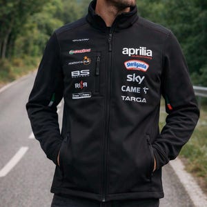 Può includere: Giacca nera con zip con diversi loghi di sponsor, tra cui "Aprilia Racing" e "Sterilgarda". La giacca ha un collo alto, due tasche anteriori e un dettaglio a strisce verdi e arancioni sulla manica. La giacca è indossata da una persona.