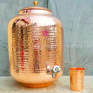 Puede incluir: Un dispensador de agua cilíndrico grande de cobre martillado con tapa metálica y grifo plateado en la base. El nombre de la marca ashtok está impreso en rosa en el frente. Un vaso de cobre martillado a juego se sitúa a la derecha del dispensador.