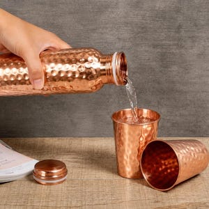 Op de afbeelding: Een gehamerde koperen waterfles schenkt water in een bijpassende gehamerde koperen beker. Een tweede lege gehamerde koperen beker ligt op zijn kant naast een koperen flesdop. De set heeft een gepolijste metalen afwerking over het geheel.
