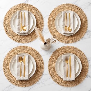 May include: A set of four round woven placemats with fringed edges, each topped with a white ceramic plate, a folded beige linen napkin, and gold-toned cutlery. A small white vase with dried pampas grass sits in the centre of the arrangement.
