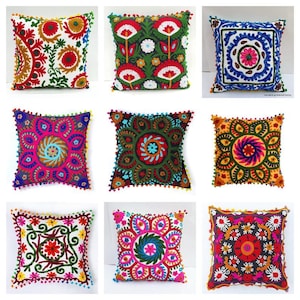 May include: Nine square decorative cushions with embroidered floral patterns. The cushions display a variety of colours, including red, green, blue, orange, and pink, with white and dark backgrounds. Each cushion has a unique design and is trimmed with small pom-poms.