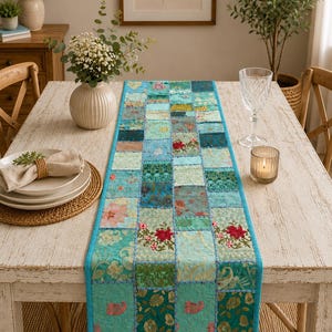 Puede incluir: Un camino de mesa rectangular con un diseño de patchwork compuesto por cuadrados de tela en tonos verde azulado, turquesa y verde. Incluye patrones florales bordados, detalles en dorado metálico y costuras azules visibles entre parches.