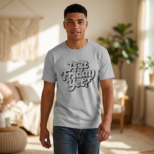 May include: A light grey t-shirt with the text "Is It Friday Yet?" in a retro font. The shirt is short-sleeved and made of a soft material. The person is wearing blue jeans.