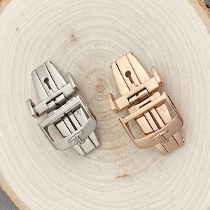 Puede incluir: Dos cierres desplegables para reloj, uno en metal plateado pulido y otro en metal color oro rosa. Cada cierre presenta un mecanismo articulado con un logotipo central en forma de T y botones laterales integrados para un cierre seguro.