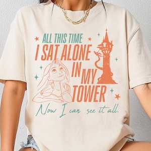 以下が含まれることがあります： 「ALL THIS TIME I SAT ALONE IN MY TOWER」のテキストと、塔とキャラクターのグラフィックが描かれたアイボリー色のTシャツ。「Now I can see it all」のテキストも含まれています。このシャツはコンフォートカラーズ素材で作られています。