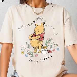 May include: Ivory-colored t-shirt with a graphic of Winnie the Pooh. The illustration shows Pooh with his arms crossed, a honey pot, and the text "I've got a rumbly in my tumbly..."