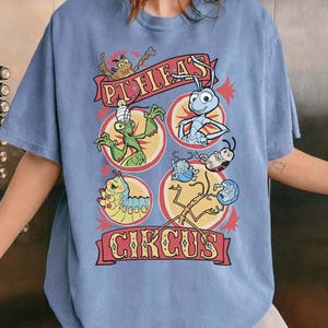 May include: Blue jean comfort colors t-shirt featuring a colorful graphic of insects in a circus setting. The design includes the text "P.T. Flea's Circus" in a banner, with various insect characters in circular frames. The shirt has a crew neck.