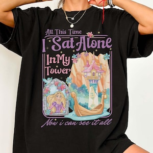 May include: Black t-shirt with a colourful graphic depicting a tower, boat, and a woman with long blonde hair. The text on the shirt reads "All This Time I Sat Alone In My Tower Now I Can See It All."