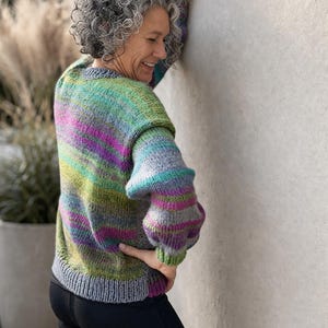 Peut inclure: Un pull tricoté à la main aux couleurs vives, avec un col rond et de longues manches bouffantes. Le pull présente des rayures horizontales vertes, violettes, roses et grises. Le bas et les poignets sont gris uni. La personne porte un pantalon noir.