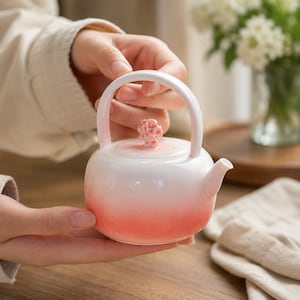 Puede incluir: Una pequeña tetera de cerámica con un cuerpo liso y redondeado que presenta un degradado de blanco a rosa coral. La tetera tiene un asa curva, un pico corto y una tapa rematada con un pequeño pomo texturizado en forma de flor coral.