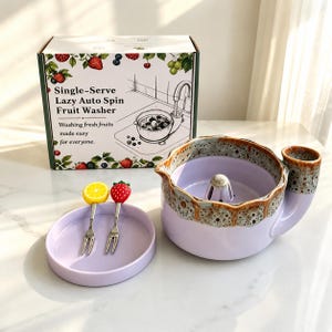 May include: A lavender ceramic fruit washing bowl with a textured brown rim, a central drainage post, and a side handle. Included are a matching lavender circular tray and two small metal forks topped with a yellow lemon and a red strawberry.