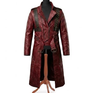 May include: A long, deep red leather coat featuring a structured collar, a front zipper closure, and two buckled straps at the waist. The garment includes a dark, textured shoulder panel adorned with metallic studs and long, fitted sleeves.