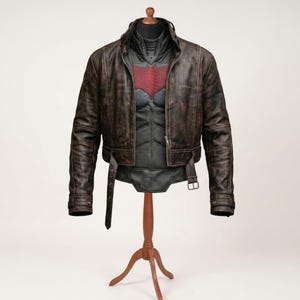 May include: A distressed brown leather jacket worn over a dark gray tactical vest featuring a prominent red bat-shaped emblem on the chest. The jacket includes a high collar, long sleeves, and a buckled belt at the waist, displayed on a wooden stand.