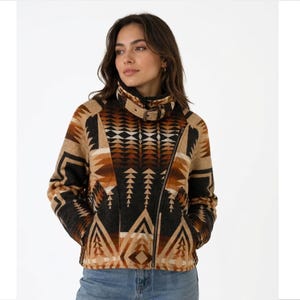 May include: A long-sleeve jacket featuring a geometric pattern in shades of black, brown, and tan. The jacket has a high collar with a buckle strap closure, an asymmetrical front zipper, and two side pockets, worn over blue denim jeans.
