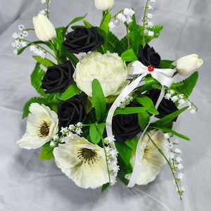 May include: A floral arrangement featuring black and white roses, white tulips, white anemones, and lily of the valley sprigs. The bouquet includes green foliage and is accented with a white ribbon tied in a bow with a small red bead at the centre.