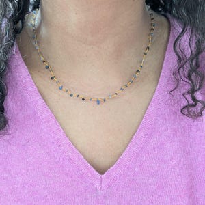 Peut inclure: Collier doré délicat orné de petites perles bleues et noires de forme irrégulière. Le collier est porté sur un pull rose à col en V. Les perles sont espacées le long de la chaîne dorée, créant un design minimaliste.