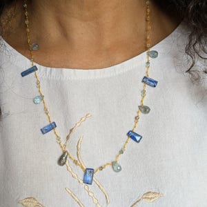 Peut inclure: Un collier délicat orné de pierres bleues rectangulaires et en forme de goutte, entrecoupées de petites perles transparentes. Les pierres précieuses sont enfilées sur une fine chaîne dorée. Le collier est présenté sur fond blanc.