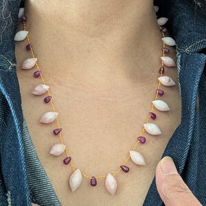 Peut inclure: Un collier délicat orné de pierres roses pâles en forme de feuilles et de petites perles rouges foncées, enfilées sur une chaîne dorée. Le collier est présenté sur une veste en jean.