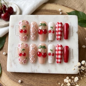 May include: A set of press-on nails with various designs. Some nails feature cherry designs with red cherries and green leaves, while others have white polka dots, red and white gingham patterns, and floral accents. The nails are displayed on a white surface.
