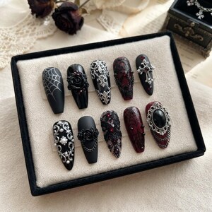 May include: A set of ten press-on nails in a coffin shape, featuring black and deep red base colors. Designs include silver filigree, black lace patterns, spider webs, 3D roses, chains, small pearls, red rhinestones, and a silver cross emblem.
