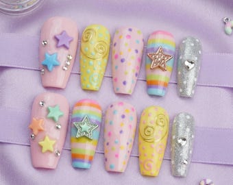 May include: A collection of ten artificial nails in various designs. The nails feature pastel pink, yellow, and rainbow colors, adorned with stars, swirls, and polka dots. Some nails have glitter or rhinestone accents, creating a playful and decorative aesthetic.