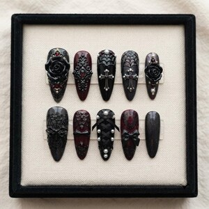 May include: A set of ten almond-shaped press-on nails in a black and deep burgundy color palette. The designs feature intricate 3D elements including black roses, silver crosses, spider webs, white pearls, lace patterns, and small black bows.