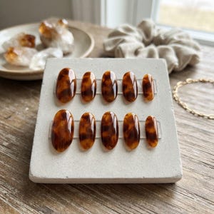 May include: A set of ten oval-shaped press-on nails featuring a glossy brown and black tortoiseshell pattern. The nails are arranged in two rows of five on a light gray rectangular display stand, showcasing varying sizes from largest to smallest.