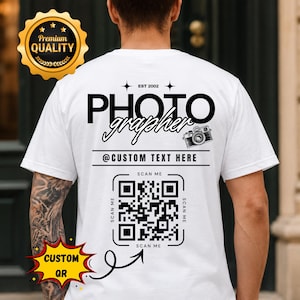 May include: A white t-shirt featuring black text on the back that reads PHOTO grapher, EST 2002, and @CUSTOM TEXT HERE. The design includes a small camera illustration and a large QR code with the text SCAN ME surrounding it on all four sides.