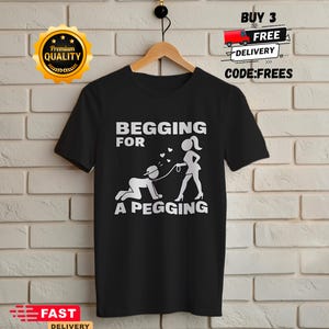 Könnte beinhalten: Ein schwarzes T-Shirt mit kurzen Ärmeln und einem weißen Grafikdruck, der eine Person auf allen Vieren mit Kappe und Halsband zeigt, die an einer Leine von einer anderen Person gehalten wird. Der Text lautet BEGGING FOR A PEGGING.