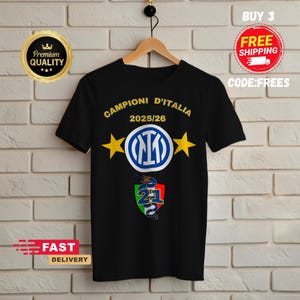 Könnte beinhalten: Schwarzes T-Shirt mit dem Text "CAMPIONI D'ITALIA 2025/26" in Gelb, zwei gelben Sternen und einem Logo. Darunter befindet sich eine Grafik mit der Nummer 21 und einem Drachen. Das T-Shirt hängt an einem Holzbügel.