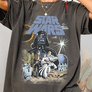Può includere: T-shirt grigio scuro con grafica di Star Wars. Il design include Darth Vader, altri personaggi e il logo di Star Wars in blu. La maglietta ha un aspetto vintage e usurato.