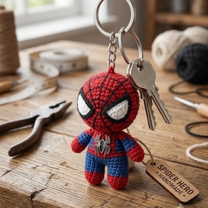 Może przedstawiać: Szydełkowany brelok Spider Hero przedstawiający czerwoną i niebieską figurkę z włóczki z dużymi białymi oczami i czarnym emblematem pająka na piersi. Figurka jest przymocowana do srebrnego metalowego kółka z dwoma kluczami i drewnianą zawieszką.