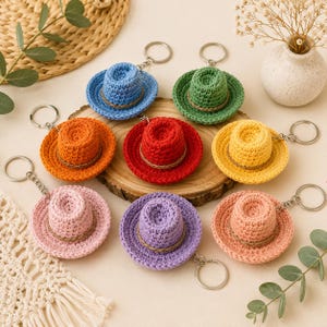 Puede incluir: Un conjunto de ocho llaveros de sombrero tejidos a ganchillo en varios colores, incluyendo azul, verde, naranja, rojo, amarillo, rosa, morado y melocotón. Cada sombrero miniatura tiene una corona texturizada, ala ancha y un cordón trenzado marrón.
