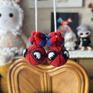 Könnte beinhalten: Zwei runde, gehäkelte Ornamente mit einem rot-blauen Spinnennetz-Muster und großen, schwarz umrandeten weißen Augen. Jede Kugel ist an einer weißen Schnur befestigt, die als Paar zum Aufhängen gedacht ist und detaillierte Garnstiche zeigt.