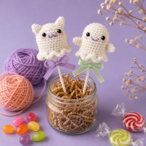 May include: Two white crocheted ghost figures on sticks, one with cat ears and a purple bow, the other with a green bow. Both have black eyes, pink cheeks, and embroidered mouths. They are placed in a glass jar filled with brown paper shreds.