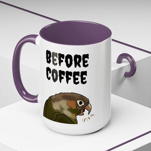 Könnte beinhalten: Ein weißer Keramikbecher mit violettem Innenraum und Henkel. Auf der Vorderseite befindet sich eine Grafik eines grün-braunen Papageis mit offenem Schnabel sowie der schwarze Schriftzug "BEFORE COFFEE" in einer fetten, ausgefransten Schriftart.