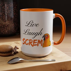 Könnte beinhalten: Weißer Keramikbecher mit orangefarbenem Innenraum und Henkel. Der Becher zeigt den Text "Live Laugh SCREM" mit einer Illustration eines Papageis. Ein Löffel und ein Macaron liegen auf einer Holzoberfläche.