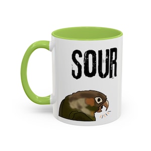Könnte beinhalten: Weiße Keramik-Tasse mit grünem Innenraum und Griff. Die Tasse zeigt das Wort "SOUR" in fetten schwarzen Buchstaben und eine Cartoon-Illustration eines Papageis mit offenem Schnabel. Der Papagei ist braun, grün und weiß.