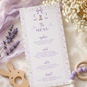 Puede incluir: Una tarjeta de menú rectangular con un borde floral de color morado claro, una ilustración central de un conejo con un lazo y el texto The MENU seguido de secciones para appetizer, salad, entrée y dessert con platos listados en tinta morada.