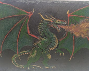 May include: A rectangular black slate plaque featuring a hand-drawn illustration of a green dragon with large wings, sharp claws, and yellow horns. The dragon is depicted in profile, breathing a cloud of orange and yellow fire from its open mouth.