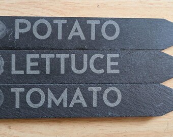May include: Three rectangular slate garden markers with pointed ends. Each marker features an engraved vegetable illustration and its corresponding name in capital letters: POTATO, LETTUCE, and TOMATO. The dark grey stone has a natural texture.
