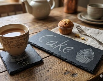 May include: A rectangular black slate serving board engraved with the word Cake and a cupcake graphic, paired with a square black slate coaster engraved with the word Tea. Both items feature natural rough edges and a dark, textured stone surface.