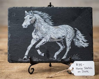 May include: A rectangular black slate plaque featuring a white hand-drawn sketch of a galloping horse. The artwork is displayed on a black metal stand. A white paper tag attached with twine reads: $35 - Horse Sketch on Slate.