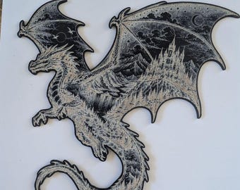 May include: A black and white wall art piece shaped like a dragon. The dragon's body features intricate stippled illustrations of a mountain landscape, a dense forest, and a large castle. The wings display a night sky with clouds and a crescent moon.