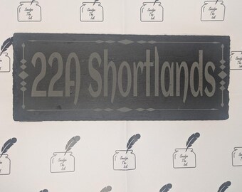 May include: A rectangular dark gray slate house sign featuring the engraved text 22A Shortlands in a bold font. The sign is bordered by a thin etched line with diamond-shaped accents on the left and right sides, creating a clean and clear display.