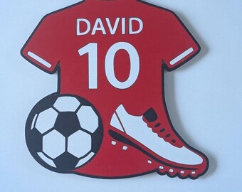 May include: A red, jersey-shaped wall decoration featuring the white text DAVID and the number 10. The design includes a black and white soccer ball graphic and a white soccer cleat with red accents positioned at the bottom of the jersey shape.