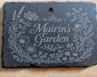 May include: A rectangular black slate plaque with a single hole at the top center. The surface features an engraved floral wreath design surrounding the text Mairin's Garden in a script font. The edges of the slate are rough and uneven.