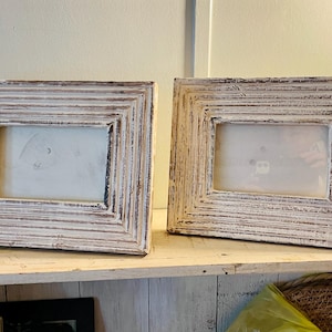May include: Two rectangular picture frames featuring a distressed white finish over a dark wood base. Each frame has a stepped, layered border design with a horizontal orientation and a clear glass insert protecting the center area.